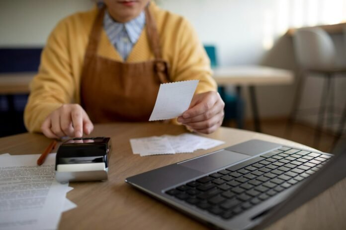 Pessoa de avental marrom e blusa amarela sentada à mesa de um restaurante, analisando recibos de papel. Em primeiro plano, uma máquina de cartão e um notebook aberto sobre a mesa de madeira, ilustrando a gestão e as estratégias de A&B para hotéis com foco em aumentar o faturamento do restaurante.