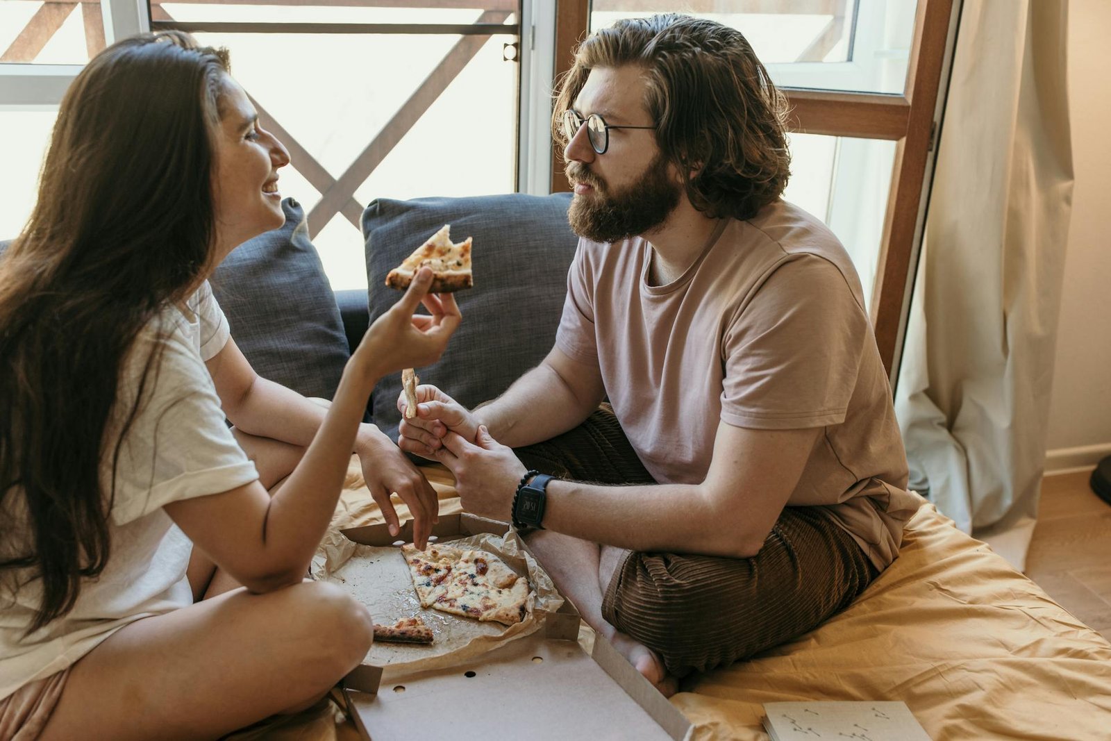 Casal sentado de pernas cruzadas em uma cama com lençol mostarda, comendo pizza de uma caixa aberta de papelão. A mulher segura um pedaço de pizza enquanto sorri para o homem de barba e óculos, ilustrando o hábito dos hóspedes de pedir delivery no quarto do hotel.