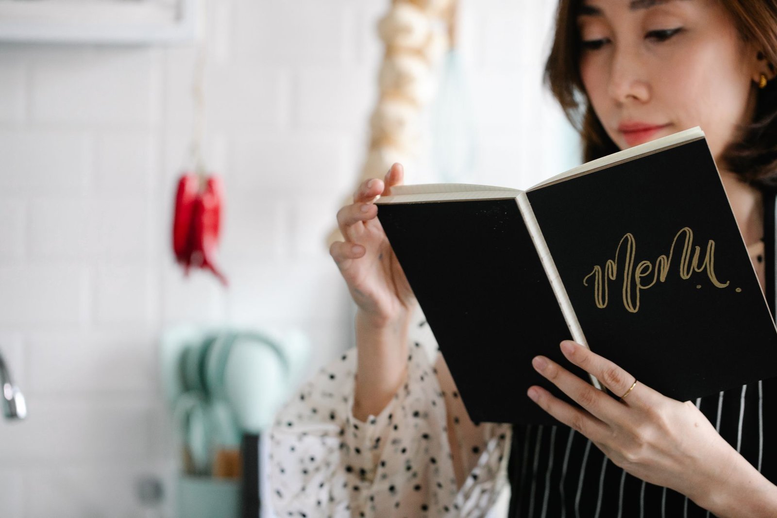 Mulher segurando e lendo um caderno preto com a palavra "Menu" escrita em dourado na capa. Ela veste um avental listrado em um ambiente de cozinha iluminado com azulejos brancos ao fundo, representando o planejamento e a engenharia de cardápio.