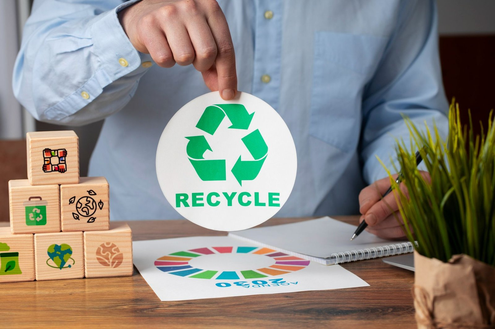 Gestor hoteleiro analisa a aplicação de boas práticas na hotelaria voltadas para a sustentabilidade. Em plano detalhe, suas mãos aparecem sobre uma mesa de madeira; uma delas segura um disco branco com o símbolo verde de reciclagem e a palavra RECYCLE, enquanto a outra segura uma caneta sobre um caderno. Ao redor há uma pirâmide de blocos de madeira com ícones sustentáveis e um infográfico impresso da Agenda 2030, ilustrando o planejamento estratégico de resíduos.