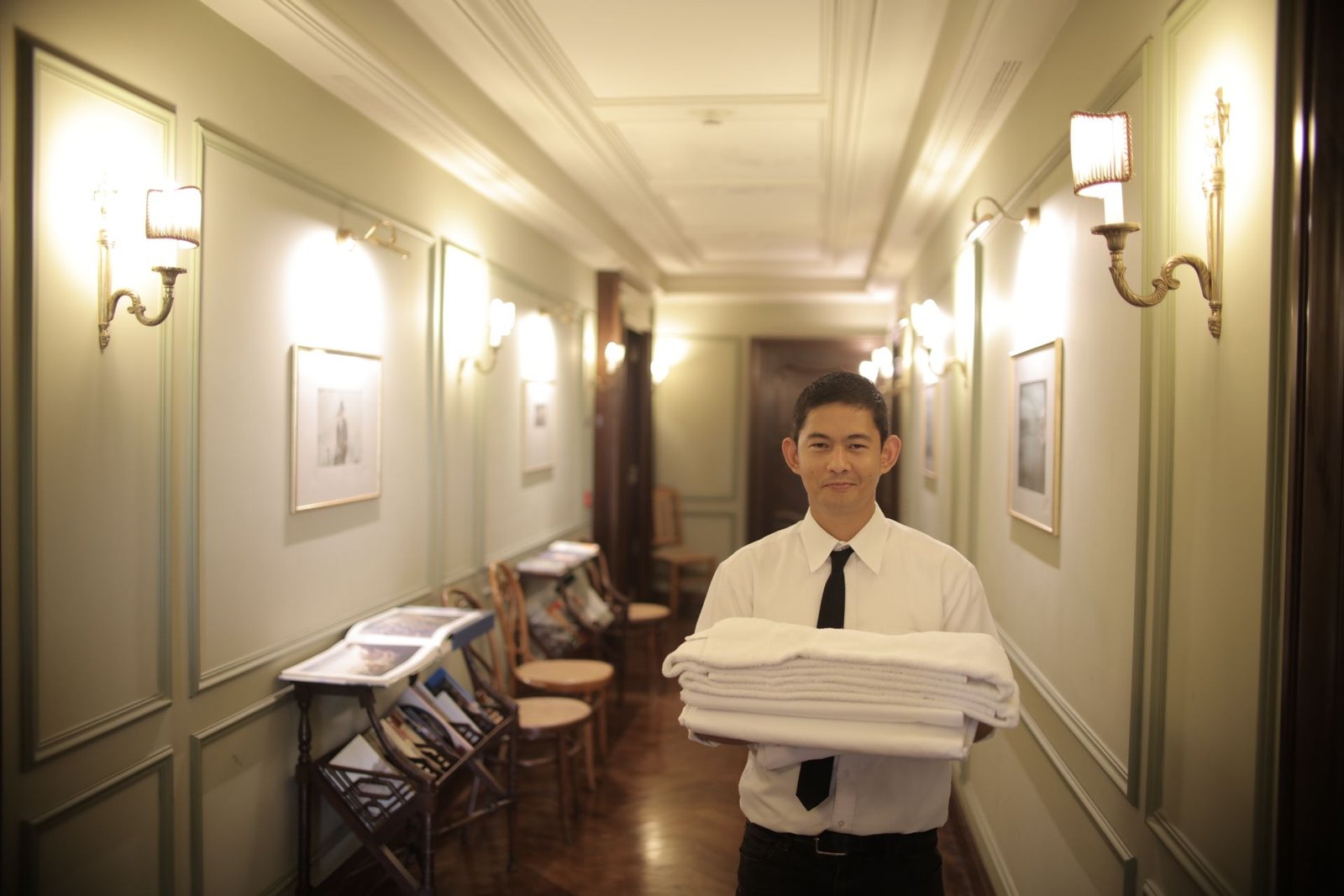 Funcionário do setor de governança sorri no centro de um corredor de hotel enquanto carrega uma pilha de lençóis e toalhas brancas limpas. Ele veste camisa social branca e gravata preta. O ambiente elegante exibe paredes em tom cinza-claro com molduras clássicas, arandelas de iluminação quente acesas e piso de madeira escura, ilustrando a rotina operacional e a conscientização sobre o uso de enxoval.