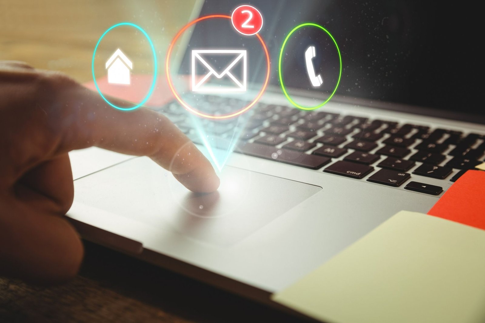 Close no dedo indicador de uma pessoa tocando o trackpad de um notebook aberto sobre uma mesa de madeira. Ícones digitais brilhantes de uma casa, um envelope com uma notificação vermelha com o número 2 e um telefone flutuam acima do teclado. No canto inferior direito, aparecem partes de post-its coloridos. A imagem simboliza a automação e o envio de e-mail marketing hiperpersonalizado na hotelaria.