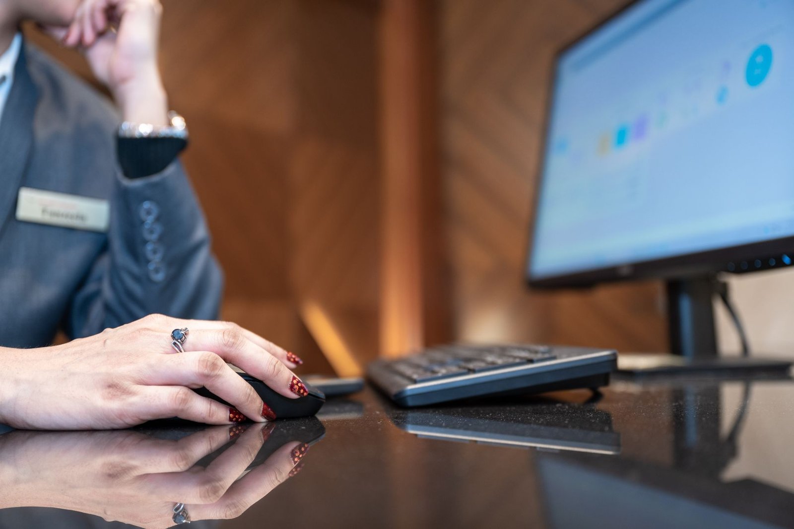 Close-up das mãos de uma recepcionista noturna com unhas pintadas de vermelho e um anel, usando um mouse de computador em um balcão de recepção preto e polido. A tela do computador exibe o gráfico de um relatório operacional. A profissional veste um terno cinza com plaqueta de identificação e o fundo da imagem aparece desfocado.