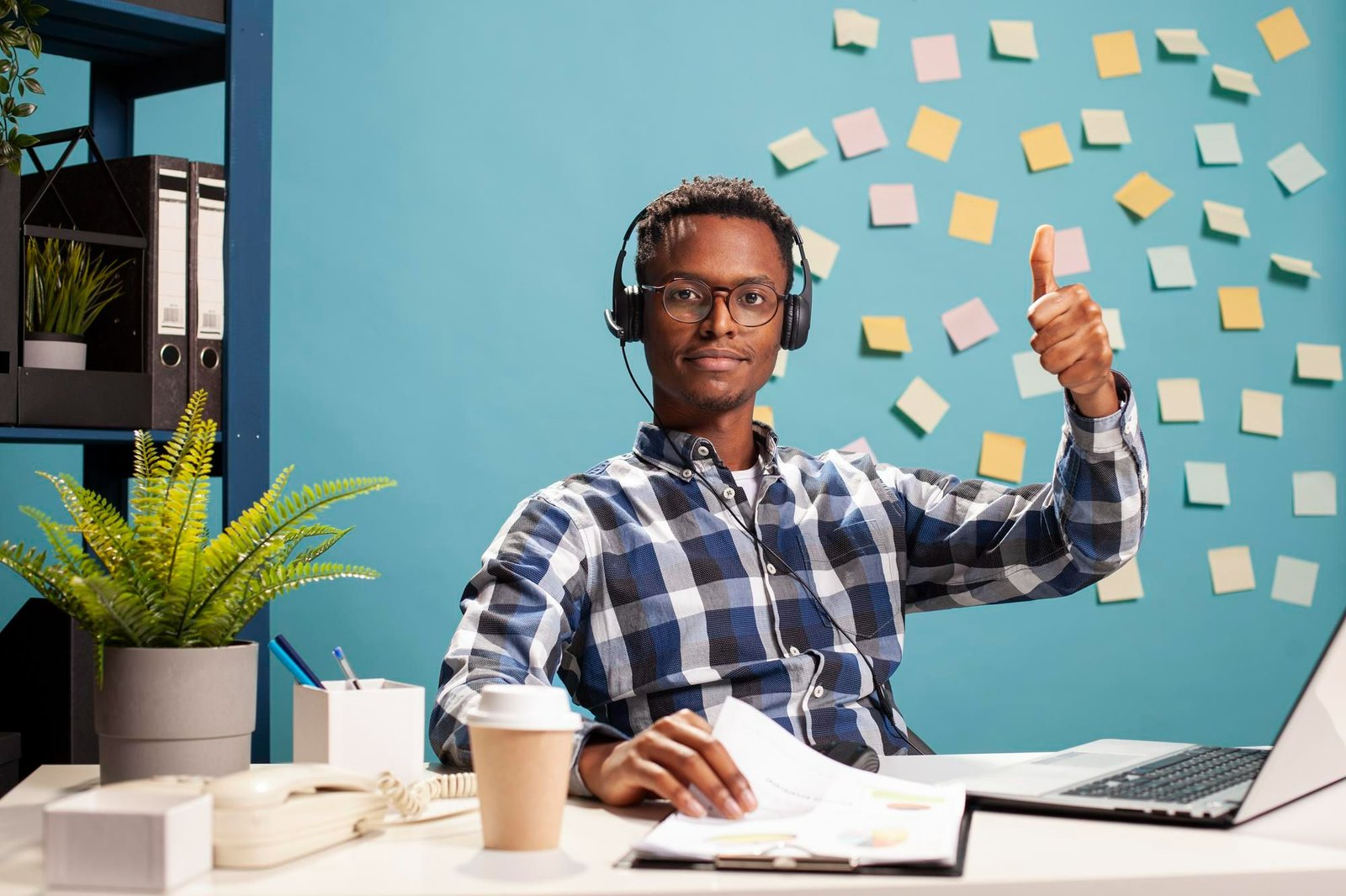 Jovem profissional negro usa óculos e headset em sua mesa de trabalho. Ele está sorrindo e fazendo um gesto de positivo com o polegar, ilustrando o sucesso nas estratégias de retenção de talentos. Ele veste camisa xadrez azul e branca, tem uma prancheta com relatórios à sua frente e está em um escritório moderno. Ao fundo, uma parede azul repleta de post-its coloridos.