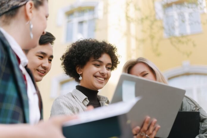 Quatro jovens profissionais sorriem e conversam enquanto olham juntos para a tela de um notebook em um ambiente externo, representando a Geração Z no mercado de trabalho.
