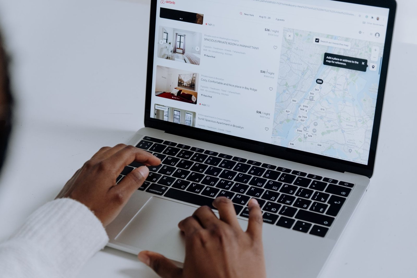 Close nas mãos de uma pessoa com pele negra digitando no teclado de um notebook prata, sobre uma mesa branca. A tela do computador exibe uma plataforma online de reserva de acomodações, mostrando uma lista de opções de hospedagem com fotos e preços do lado esquerdo, e um mapa interativo de localização do lado direito, ilustrando a facilidade e a autonomia do usuário ao realizar pesquisas e reservas online de hotéis.