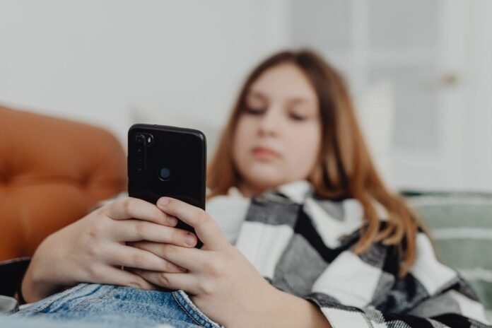 Close nas mãos de uma mulher deitada no sofá, com expressão de tédio, enquanto segura um celular preto. O rosto e o corpo estão desfocados ao fundo. Imagem contextualiza o cansaço e a fadiga digital.