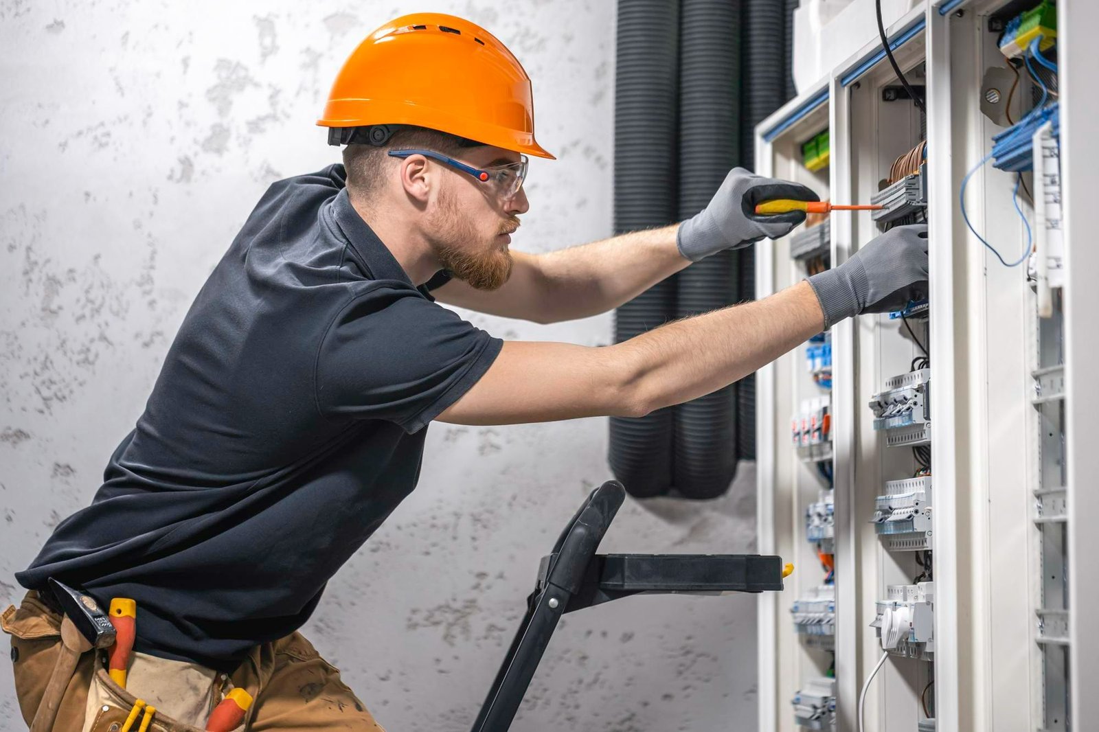 Técnico de capacete e óculos de proteção utiliza chaves de fenda para ajustar fiação em painel elétrico aberto. A imagem realça a complexidade do trabalho, motivo pelo qual é preciso reduzir custos de manutenção de forma estratégica em hotéis.