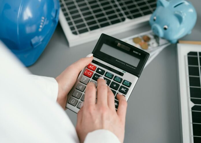 Mãos de uma pessoa utilizando uma calculadora sobre uma mesa de trabalho com planilhas, moedas, um cofrinho de porquinho azul e um capacete também azul, que configura um EPI (Equipamento de Proteção Individual). A imagem representa o cálculo estratégico necessário para planejar e reduzir custos de manutenção em hotéis.