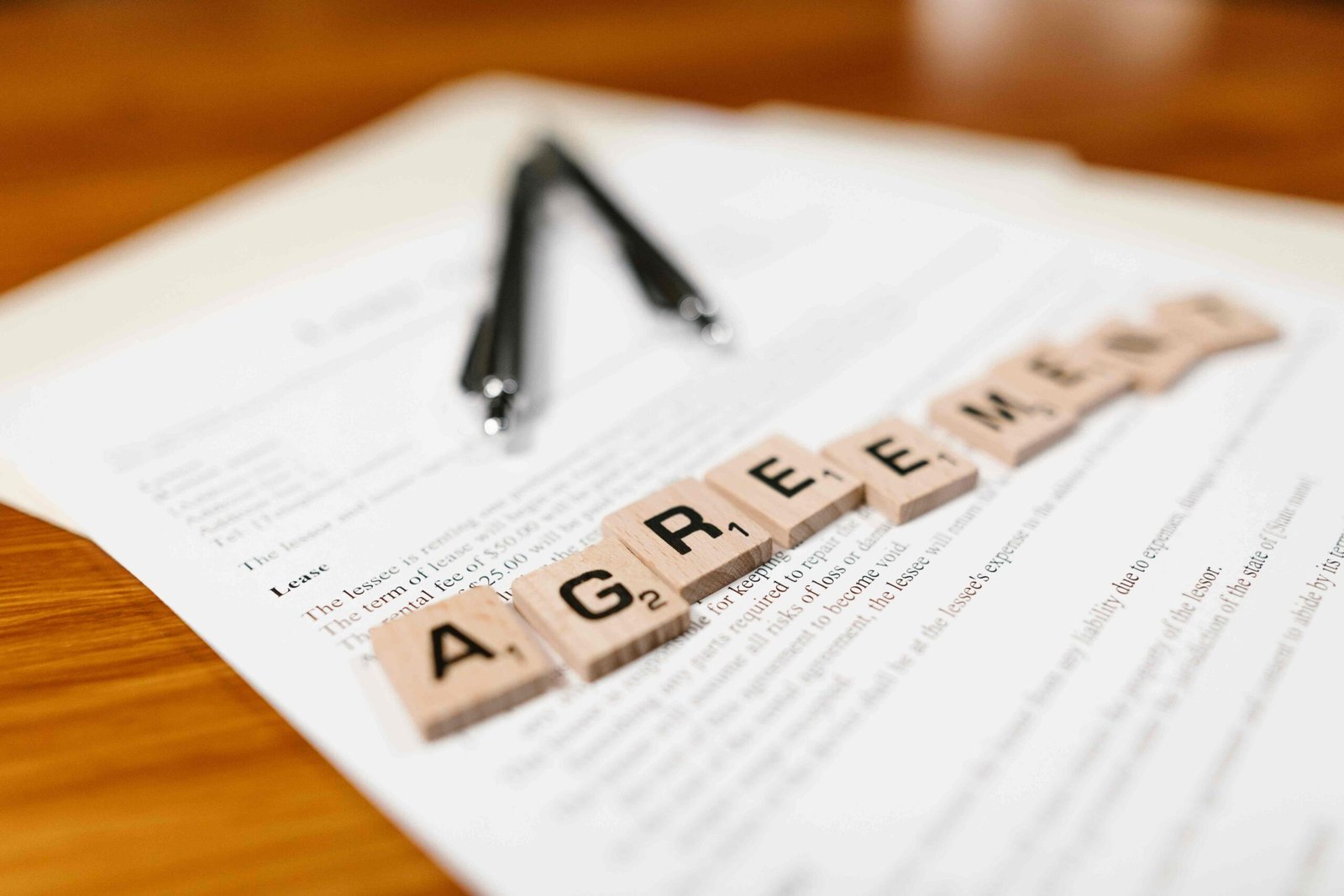 Blocos de madeira com letras formam a palavra "AGREEMENT" (Acordo) sobre um contrato impresso com duas canetas pretas ao fundo. A imagem ilustra a formalização de Acordos de Nível de Serviço, os SLAs, na gestão de fornecedores de hotéis.