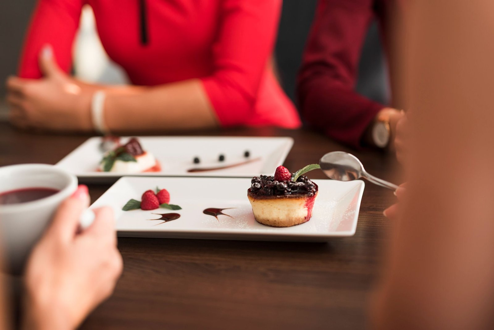 Uma pessoa segura uma colher prestes a saborear uma mini cheesecake de frutas vermelhas decorada com uma folha de hortelã. O prato retangular branco traz ainda calda de chocolate e framboesas frescas. Ao fundo, clientes aparecem desfocados conversando ao redor da mesa de madeira.