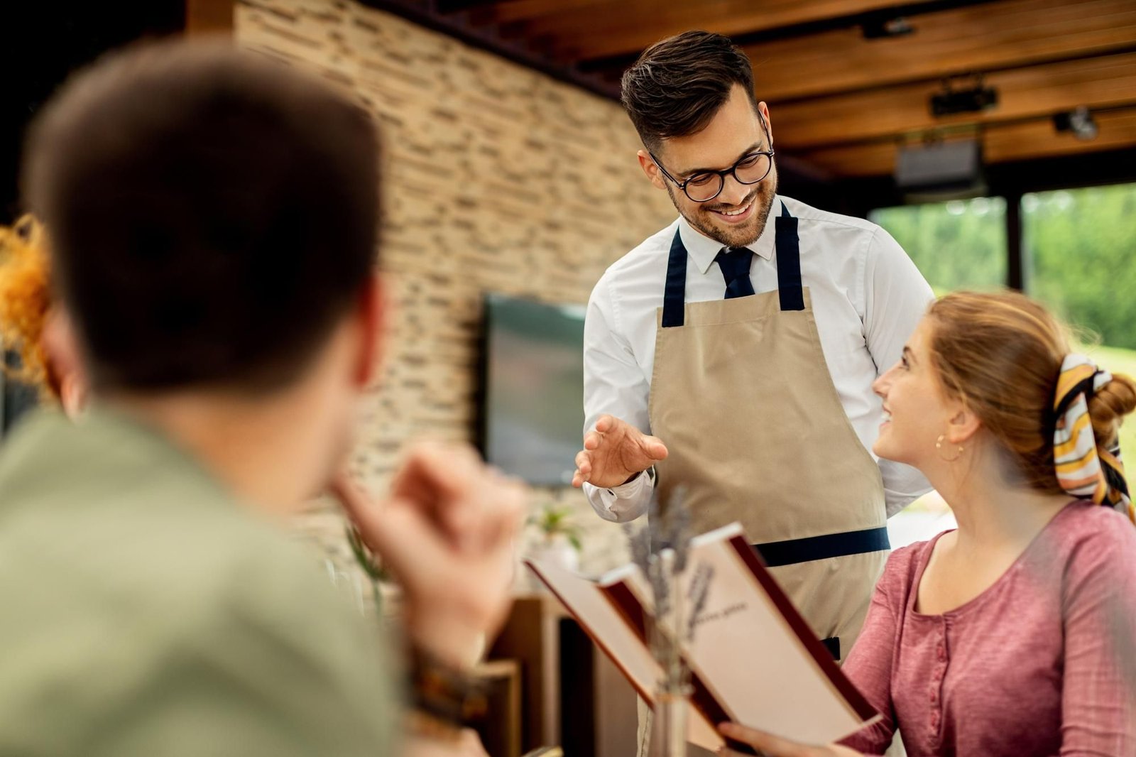 Um garçom sorridente, vestindo camisa social, gravata e avental de algodão, conversa com uma cliente sentada à mesa em um restaurante de hotel bem iluminado. O garçom gesticula levemente com a mão esquerda enquanto explica o cardápio, que está aberto nas mãos da cliente. Ela olha para ele, atenta e sorrindo. Outros clientes estão sentados à mesa, em primeiro plano, desfocados. O ambiente é acolhedor, com uma parede de pedra texturizada e vegetação visível através de uma grande janela ao fundo.