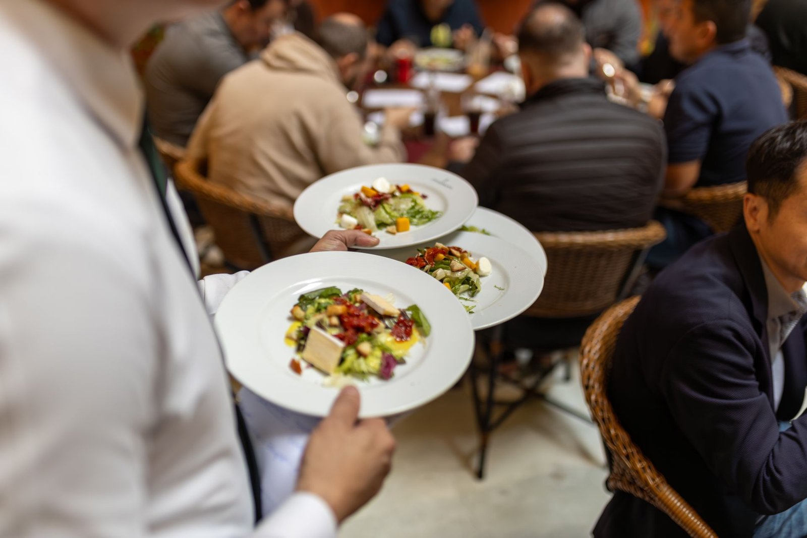 Em primeiro plano, um garçom vestindo camisa social branca carrega habilidosamente três pratos de salada com queijo e tomates secos em direção a uma grande mesa redonda. Ao fundo, um grupo de clientes aparece desfocado, conversando e jantando sentados em cadeiras de vime. A imagem ilustra a agilidade e a dinâmica do atendimento em restaurante de hotel.