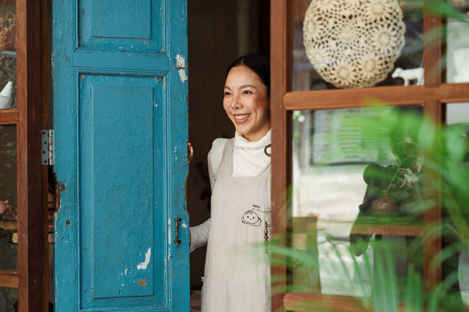 Mulher sorridente usando avental claro abre uma porta rústica de madeira azul, demonstrando uma recepção calorosa aos clientes na entrada do estabelecimento. A cena reflete a hospitalidade e o cuidado no atendimento em restaurante de hotel.