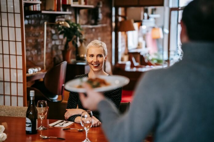 Uma mulher loira com cabelo curto e um vestido preto de bolinhas está sentada em uma mesa em um restaurante de hotel, sorrindo enquanto um garçom, visto de costas em primeiro plano, segura um prato de comida em sua direção. A mesa está posta com uma garrafa de vinho, duas taças de vinho e talheres. O fundo é um espaço de restaurante aconchegante e bem decorado, com paredes de tijolos, estantes e uma divisória de estilo japonês.