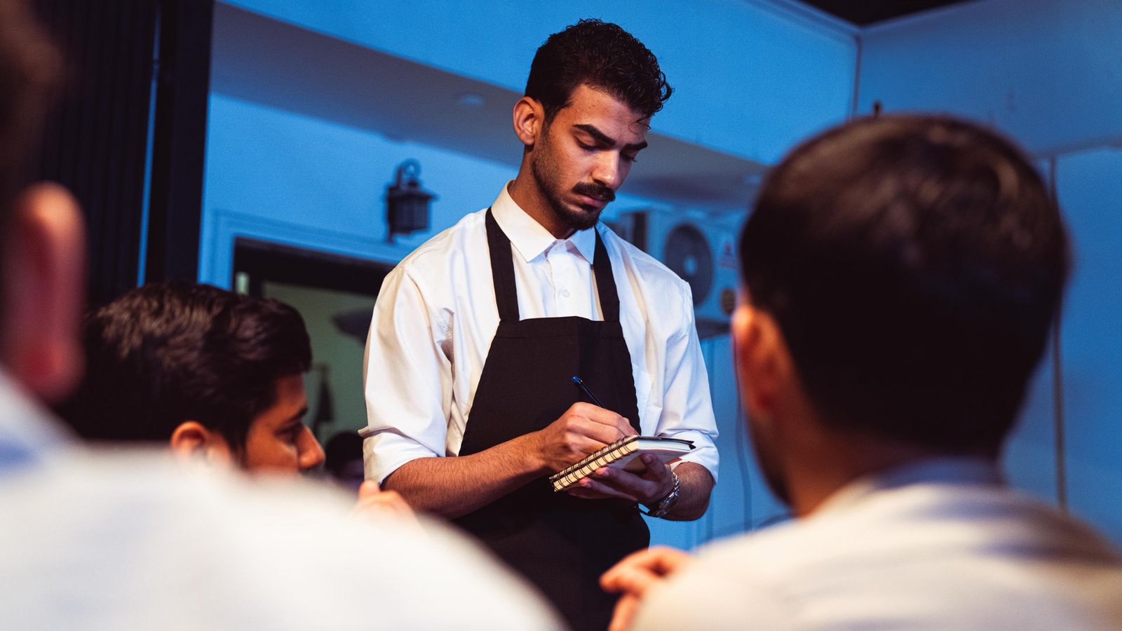 Um garçom concentrado, vestindo camisa social branca e avental preto, anota um pedido em um pequeno bloco de notas espiral. Em primeiro plano e desfocados, os clientes são vistos de perfil e de costas. O ambiente ao fundo apresenta uma iluminação suave com tons azulados, destacando o momento do atendimento em restaurante de hotel.
