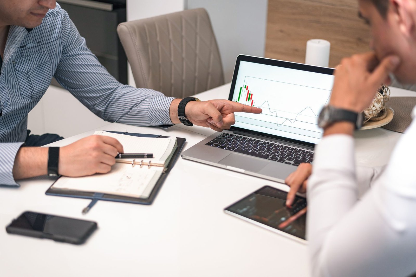 Reunião de equipe de gestão hoteleira para análise de KPIs. Um gerente aponta para um gráfico de desempenho financeiro (como GOPPAR ou RevPAR) na tela do laptop, enquanto outro consultor analisa dados complementares no tablet, refletindo sobre estratégias de otimização de margem de lucro. A cena inclui uma agenda de anotações e smartphone sobre a mesa.