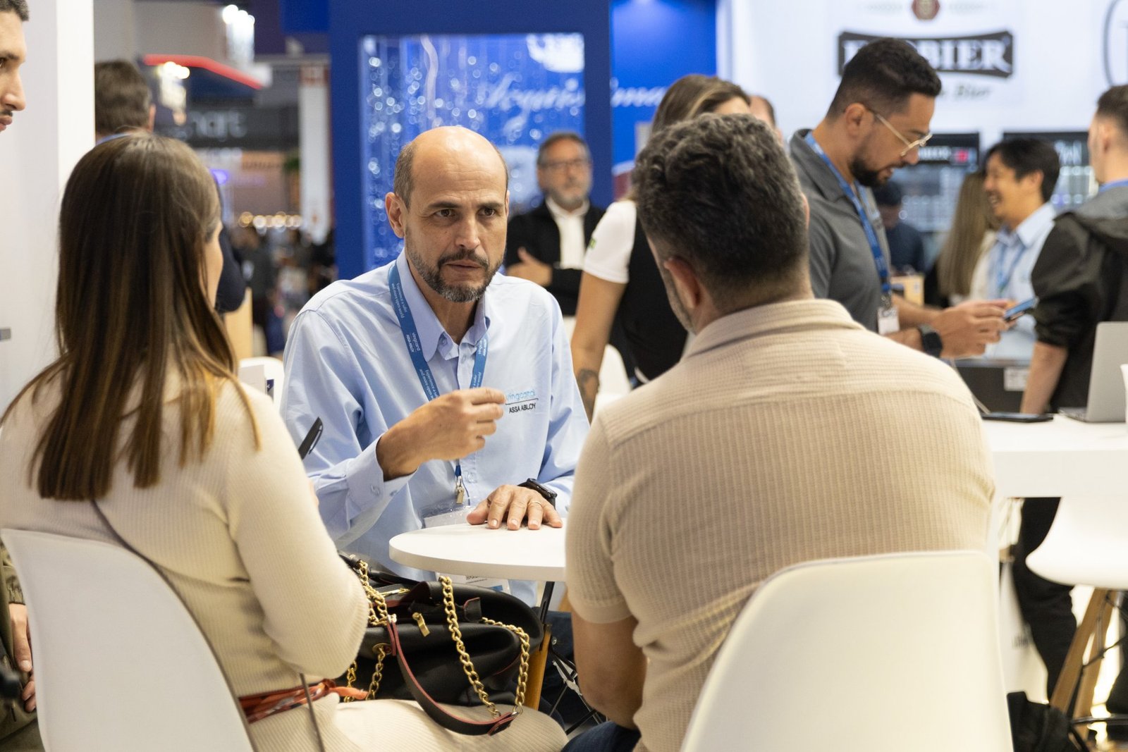 Três pessoas sentadas ao redor de uma pequena mesa em um estande. Um homem de camisa social azul-clara gesticula concentrado enquanto se dirige a dois visitantes sentados de costas para a câmera, conduzindo uma conversa de negócios.