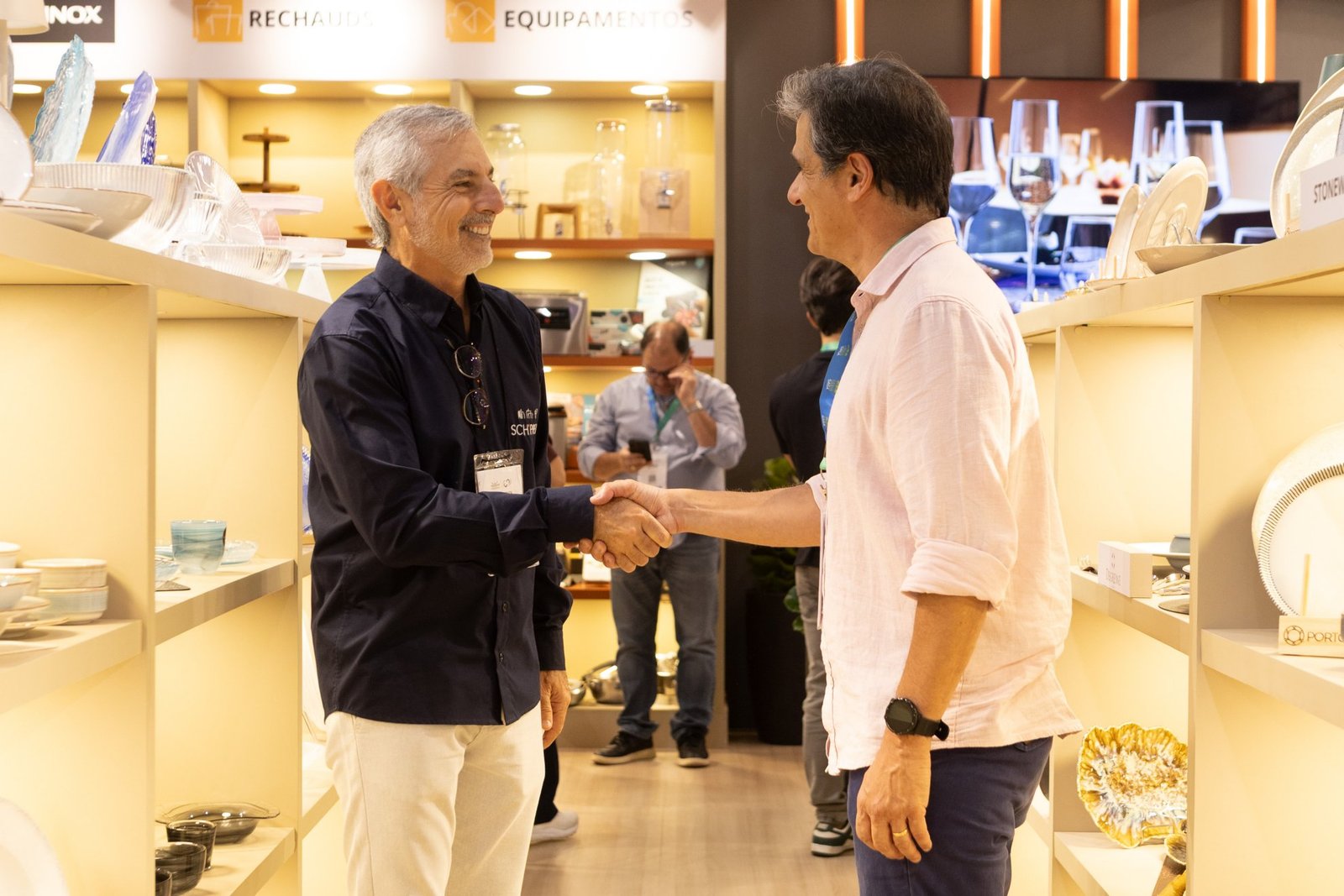 Dois executivos sorrindo apertam as mãos, aparentemente selando um acordo no corredor de um estande na Equipotel 2025.