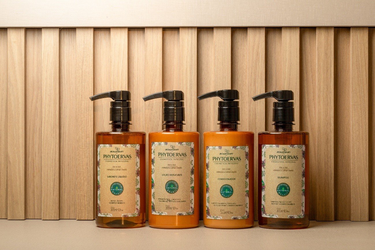 Quatro frascos dispensers com pump da marca Phytoervas alinhados lado a lado: sabonete líquido, loção hidratante, condicionador e shampoo. Os frascos possuem rótulos bege com detalhes florais e estão dispostos à frente de um painel ripado de madeira clara.