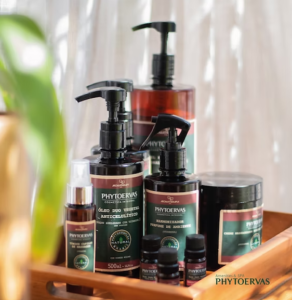 Linha de produtos da Phytoervas, uma das principais fornecedoras de amenities para hotéis, disposta em uma bandeja de madeira. A imagem mostra embalagens escuras com rótulos nas cores verde e vinho, incluindo frascos de dispenser com pump, tônico, perfumador de ambiente em spray, pote de creme e óleos essenciais.