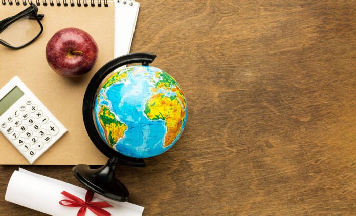 Elementos sobre mesa de madeira simbolizam o debate sobre a profissão de turismólogo. Um globo terrestre de mesa aparece ao lado de um diploma com fita vermelha, calculadora e caderno, representando a formação acadêmica e o mercado de trabalho na área.