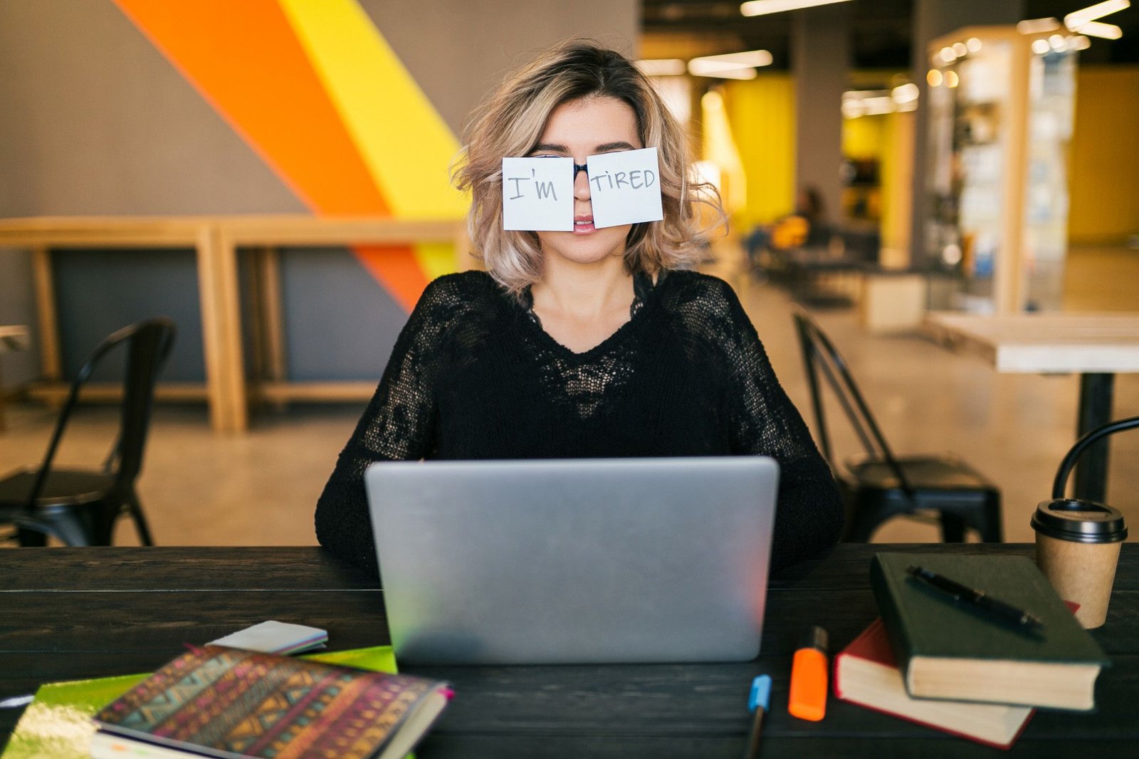 Uma jovem sentada diante de um notebook em um ambiente de escritório, com dois papéis adesivos colados sobre os olhos onde se lê "I'm" ("eu estou") e "Tired"("cansada"). A imagem ilustra o esgotamento profissional e a exaustão que podem comprometer a qualidade do atendimento na recepção do hotel.