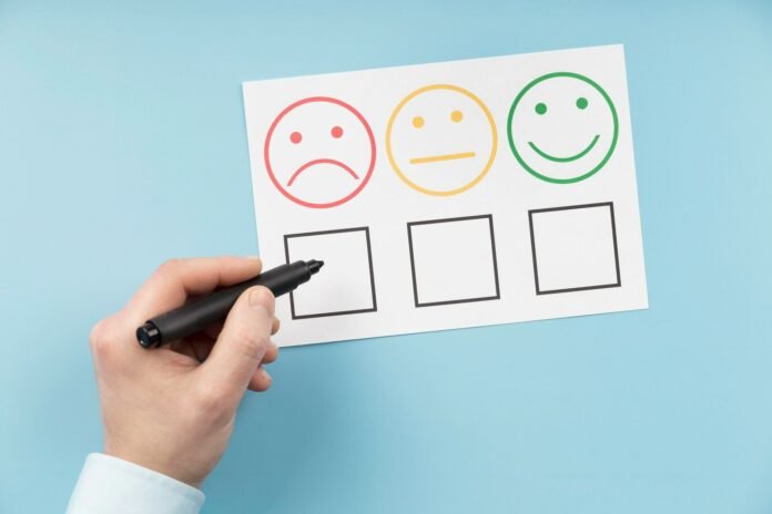 Uma mão segura uma caneta preta sobre um papel de avaliação de atendimento com três emoticons: um triste (vermelho), um neutro (amarelo) e um feliz (verde). A caneta está apontada para o quadrado abaixo do emoticon triste, sinalizando insatisfação e reclamações dos hóspedes.