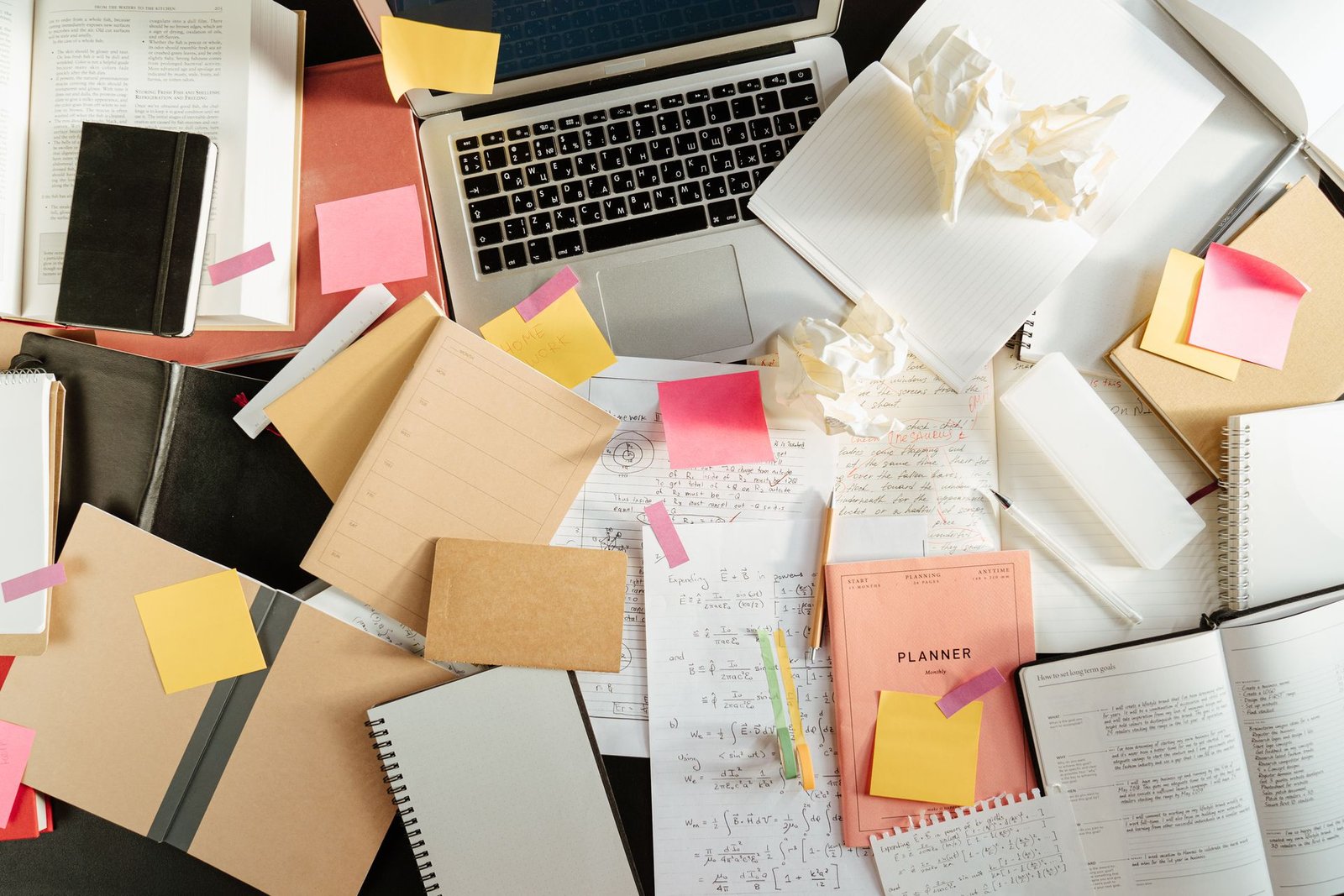 Visão aérea de uma mesa de trabalho completamente desorganizada e cheia de objetos espalhados. Há um notebook aberto cercado por diversas cadernetas, livros abertos, folhas com anotações e fórmulas matemáticas, canetas, papéis amassados e muitos blocos de notas adesivas coloridas (nas cores rosa e amarelo) colados por toda a parte, ilustrando o caos operacional e a sobrecarga na rotina de processos.