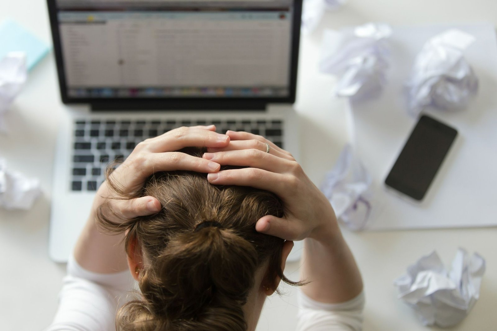 Vista superior de uma pessoa com as mãos na cabeça, demonstrando frustração em frente a um notebook. Ao redor há papéis amassados e um celular sobre a mesa. A imagem representa a dificuldade e o estresse da equipe de Compras quando a comunicação com outras áreas não é eficiente, inviabilizando o controle de estoque.