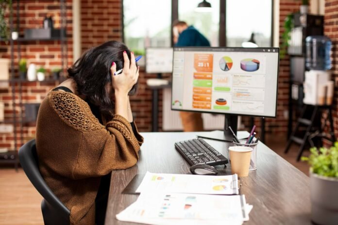 Foto de uma mulher de cabelo curto e casaco marrom, sentada em uma mesa de escritório com a cabeça entre as mãos, transmitindo desespero e preocupação. Diante dela, um monitor de computador exibe gráficos de pizza e dados financeiros, representando a análise do impacto financeiro do aumento das comissões da Booking nos hotéis. A mesa tem um teclado, mouse e relatórios impressos. O ambiente é um escritório moderno com paredes de tijolos expostos.