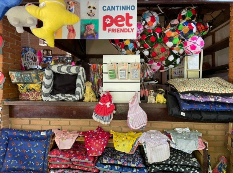 Foto em plano médio de uma seção de loja de presentes com produtos para animais de estimação. Sobre uma prateleira de madeira escura e sob uma parede de tijolos à vista, há uma placa suspensa com a frase "CANTINHO PET FRIENDLY" e fotos de cães. O espaço está repleto de itens coloridos, incluindo camas para cães de vários tecidos e estampas, brinquedos de pelúcia (como uma grande estrela amarela), bolinhas coloridas em uma rede, roupinhas penduradas e dobradas em prateleiras inferiores, guias e estatuetas decorativas de cães. O ambiente é acolhedor e cheio de detalhes, ilustrando a oferta de produtos para hóspedes com animais na Rede dos Sonhos.