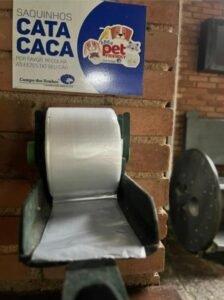 Foto em close de um dispensador de saquinhos plásticos brancos para recolhimento de fezes de cães, fixado em uma mureta de tijolos. Acima dele, uma placa azul e branca traz o aviso "Saquinhos cata caca - Por favor, recolha as fezes do seu cão", com a identidade visual de área pet friendly do hotel Campo dos Sonhos.