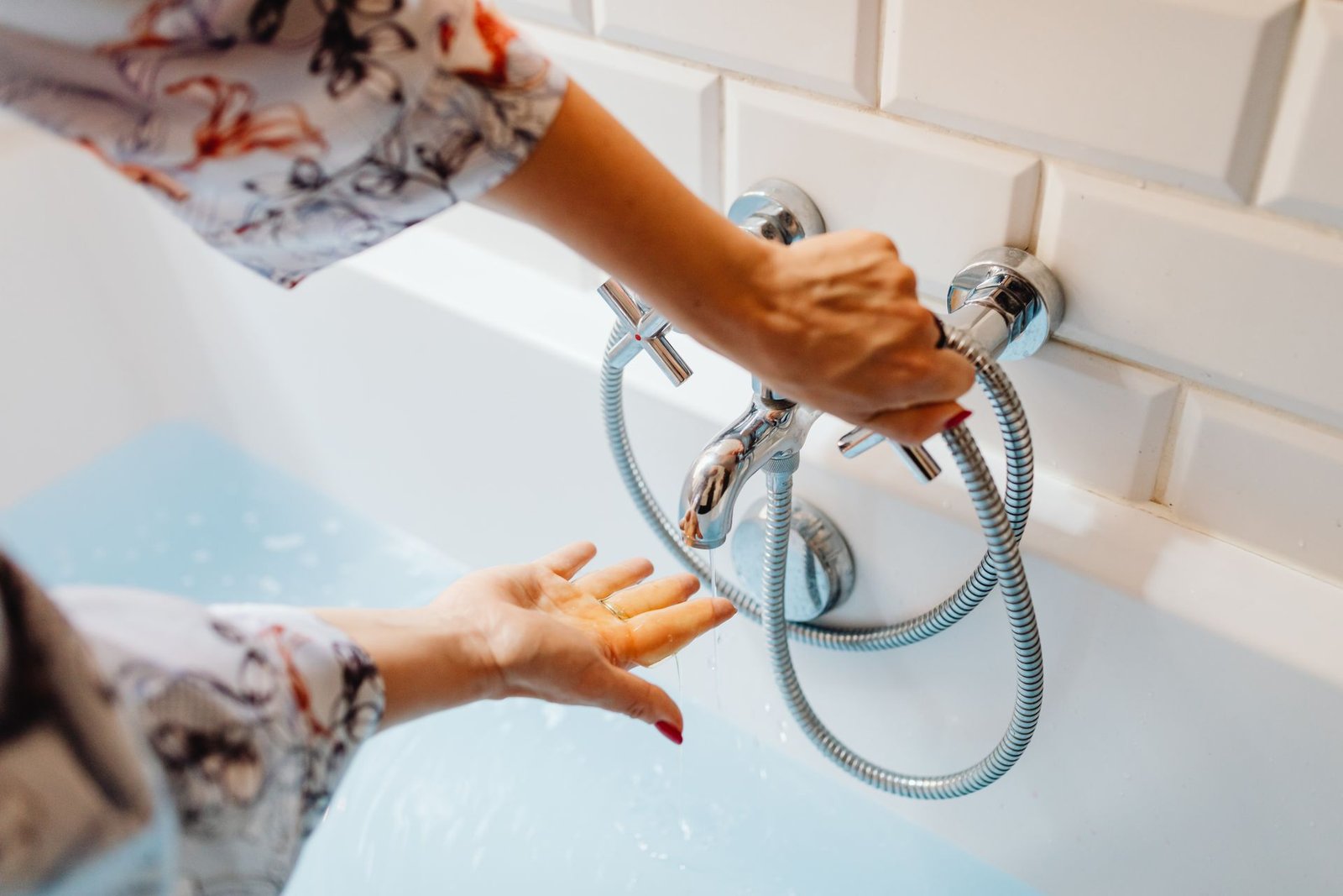Close das mãos de uma pessoa ajustando o misturador e testando a água fria que sai da torneira da banheira, ilustrando a falha no aquecimento do chuveiro devido à falta de manutenção.