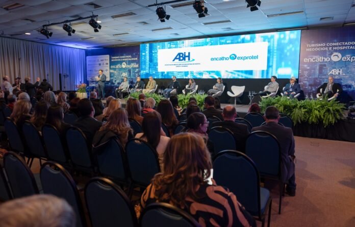Auditório com centenas de pessoas assistindo a um painel de discussão no palco principal. Os telões de LED mostram as logomarcas da ABIH-SC e do evento Encatho & Exprotel, com as frases 