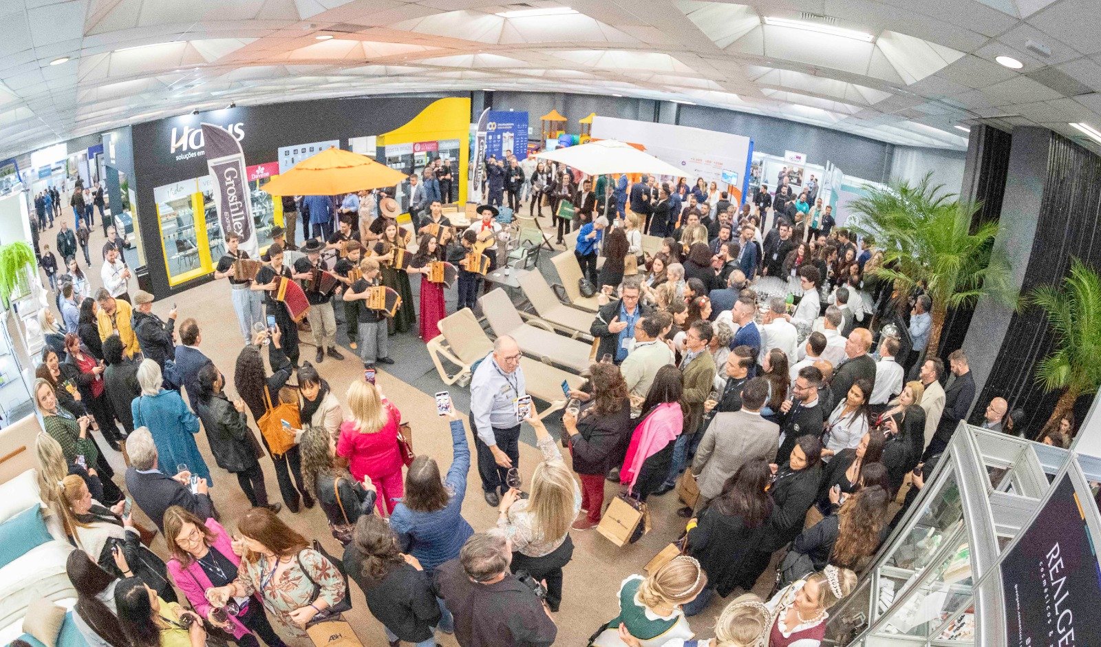 Vista panorâmica de um grande pavilhão da feira de negócios Exprotel, edição de 2025. A imagem captura centenas de pessoas participando de atividades de networking, assistindo a uma apresentação musical ao vivo e visitando os estandes de expositores.