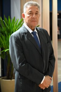 Retrato fotográfico de Alexandre Sampaio, um homem de pele clara e cabelos curtos e grisalhos, em pé, do peito para cima. Ele veste um terno cinza-escuro, camisa branca e gravata azul-marinho com pequenos padrões brancos. Ele está com as mãos entrelaçadas à frente do corpo e olha diretamente para a câmera com uma expressão séria e profissional. O fundo é um ambiente interno moderno e desfocado, com uma planta verde em um vaso à esquerda e divisórias de madeira à direita.