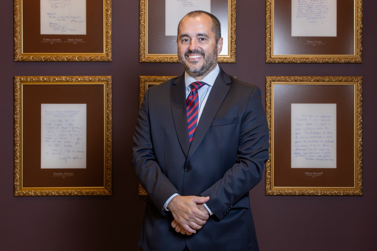 Retrato oficial de Elber Alves Justo, presidente da Associação Comercial de Santos. Ele veste terno e gravata e sorri, com as mãos cruzadas. O fundo exibe uma parede com documentos históricos emoldurados em dourado.