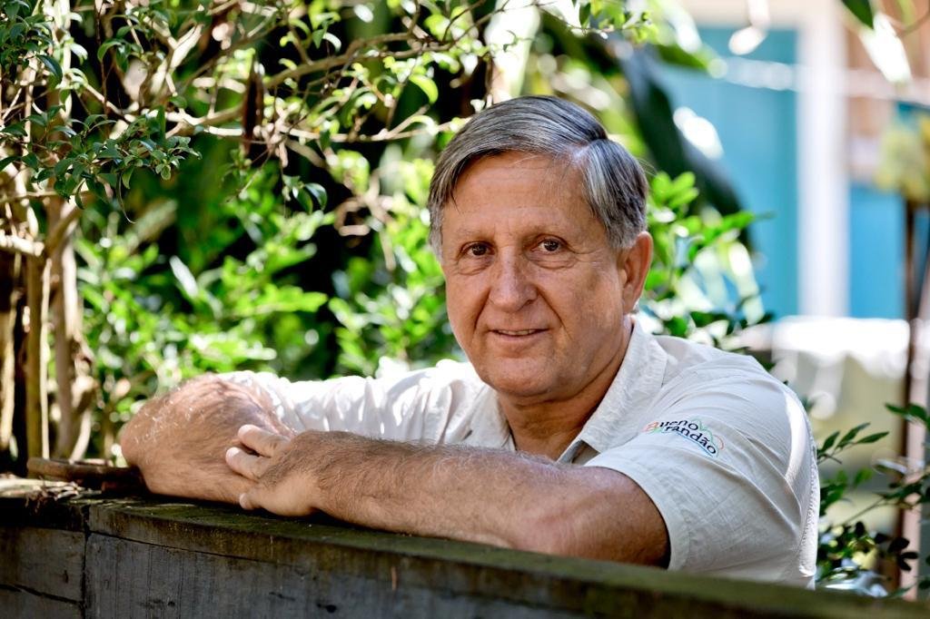 Retrato de José Fernandes, diretor da Rede dos Sonhos, em ambiente externo com fundo de folhagens verdes iluminadas pelo sol. Ele é um homem de meia-idade, cabelos grisalhos curtos, sorri levemente e veste uma camisa polo de manga curta. Ele está apoiado com os braços cruzados sobre uma mureta de madeira rústica escura, olhando para a câmera.