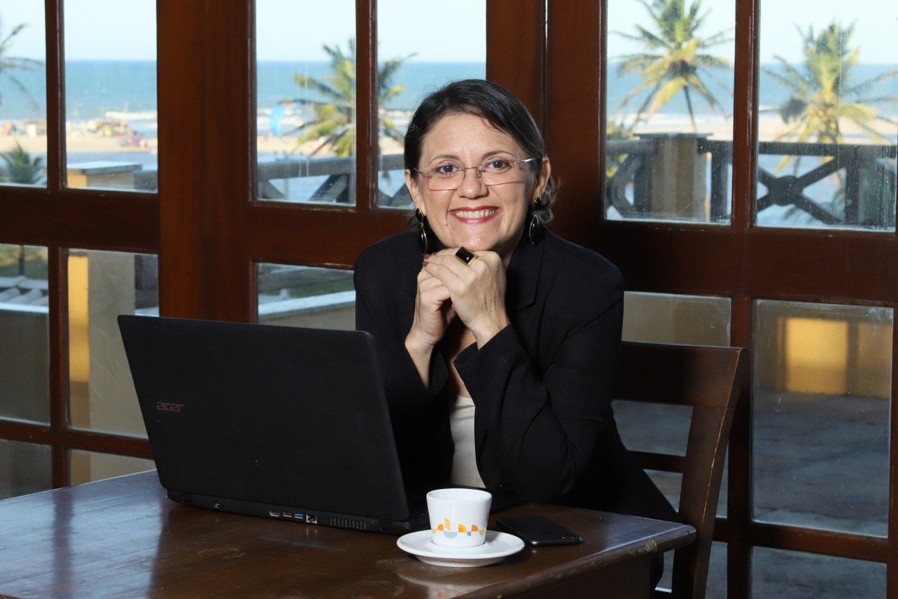 Retrato de Silvia Guimarães, uma mulher branca, de cabelos pretos curtos, usando óculos de grau e um blazer preto sobre blusa clara, sorrindo sentada à mesa. Ela está com as mãos apoiadas no queixo e olha para a câmera. Diante dela, estão um notebook preto aberto, uma xícara de café e um celular. Ao fundo, grandes janelas de madeira mostram uma vista externa com coqueiros e praia, contextualizando o debate sobre o mercado de turismo e hotelaria.