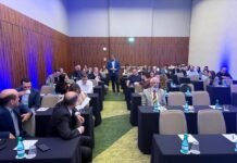 ABIH-SP orienta hotelaria sobre os impactos da reforma tributária e novas práticas de precificação Vista geral de uma sala de conferência em formato de auditório, com paredes de madeira e iluminação azul cênica. Um palestrante com blazer azul está de pé no corredor central, fazendo uma apresentação sobre os impactos da reforma tributária para profissionais do setor hoteleiro, sentados em mesas de reunião.