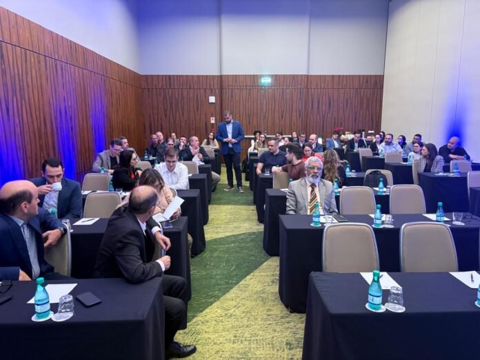 Vista geral de uma sala de conferência em formato de auditório, com paredes de madeira e iluminação azul cênica. Um palestrante com blazer azul está de pé no corredor central, fazendo uma apresentação sobre os impactos da reforma tributária para profissionais do setor hoteleiro, sentados em mesas de reunião.