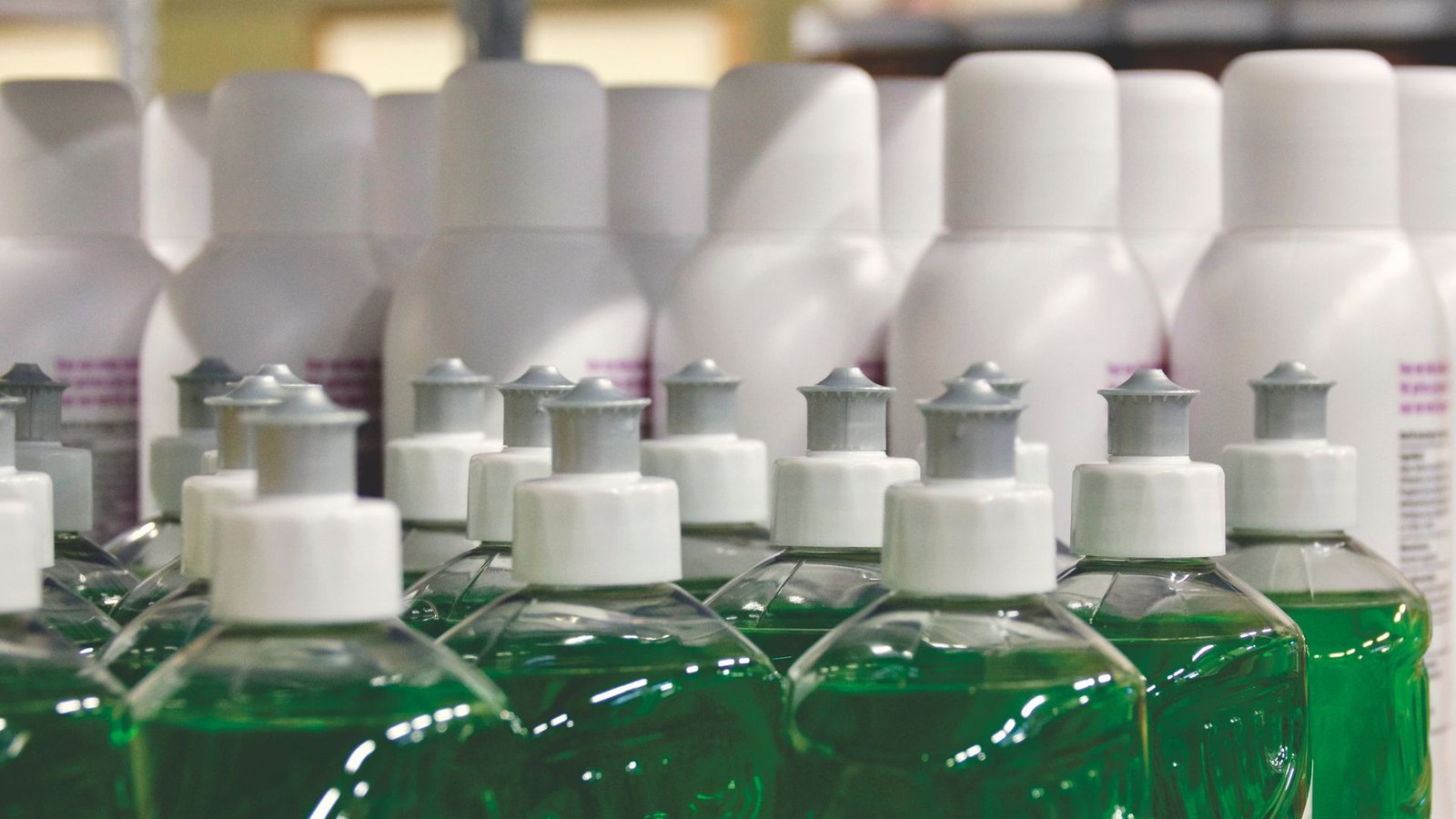 Fileiras simétricas de embalagens de produtos de limpeza organizadas em uma prateleira. Em primeiro plano, frascos transparentes com líquido verde e tampas brancas aparecem alinhados e, ao fundo, frascos plásticos inteiramente brancos estão separados, ilustrando a necessidade de classificação dos produtos em categorias para a organização do estoque.