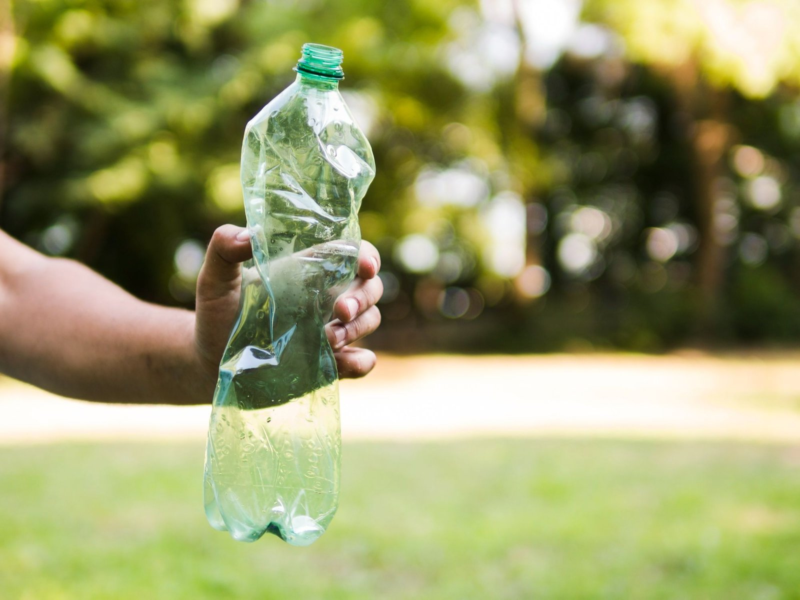 Mão segurando uma garrafa plástica PET transparente e amassada contra um fundo verde de árvores desfocadas. A imagem ilustra o impacto ambiental dos plásticos descartáveis de uso único e a importância da redução de plástico em hotéis.