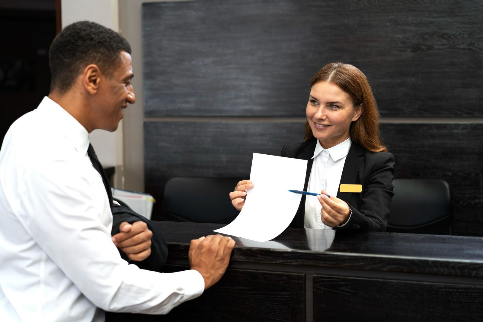 Recepcionista de hotel sorri enquanto oferece uma folha de papel e uma caneta para um hóspede no balcão. Aderir à comunicação escrita é uma das maneiras de demonstrar acessibilidade na recepção do hotel. 