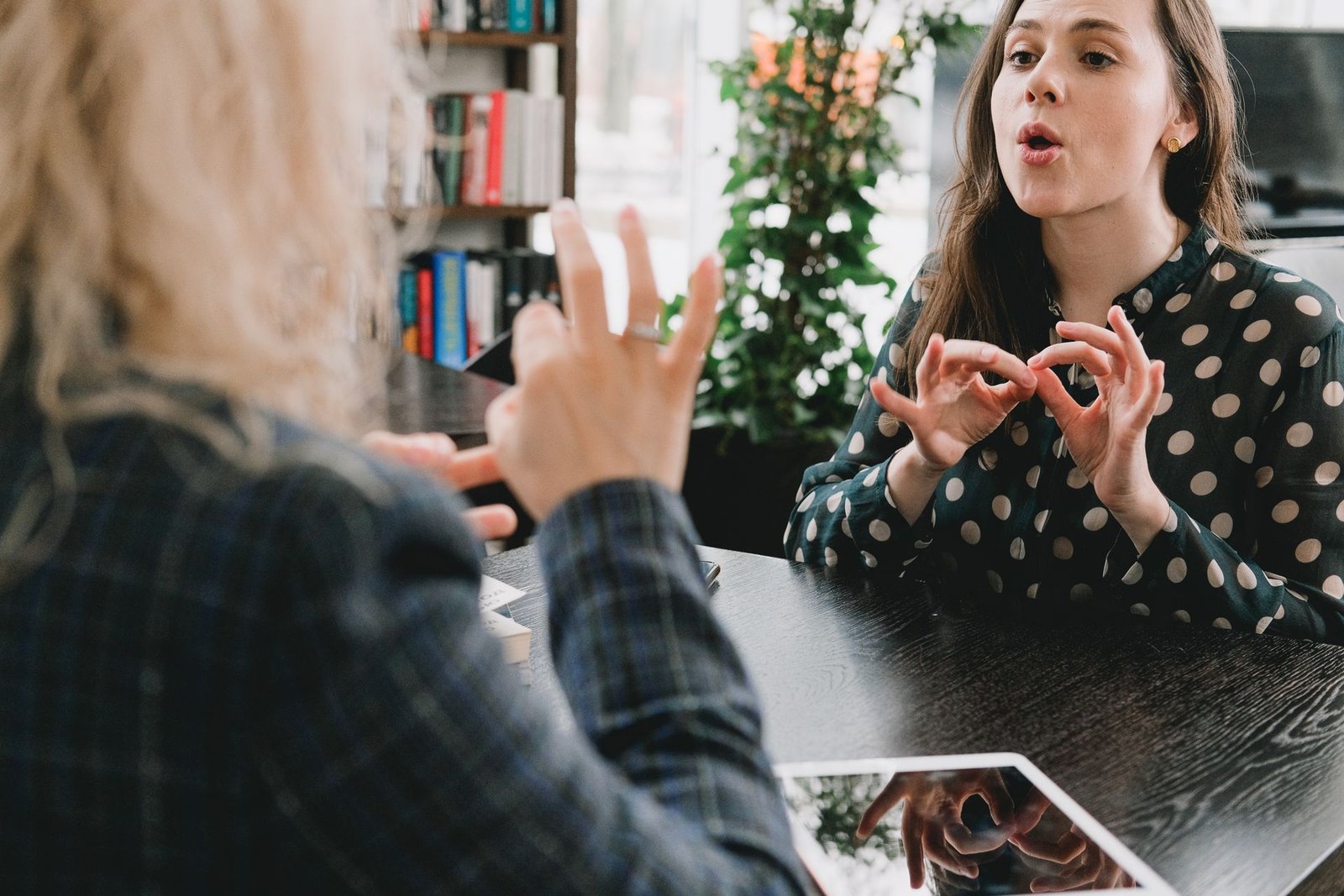Uma mulher com blusa de bolinhas (à direita) sinaliza com as duas mãos e expressão facial ativa, olhando para uma pessoa loira (à esquerda, de costas). Elas estão em um ambiente interno com livros ao fundo, conversando em linguagem de sinais.