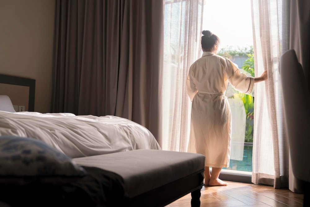 Mulher de costas e vestindo um roupão enquanto abre as cortinas de um quarto de hotel iluminado pela luz da manhã, ilustrando os pequenos detalhes sensoriais e o prazer proporcionado pela vista ao acordar.