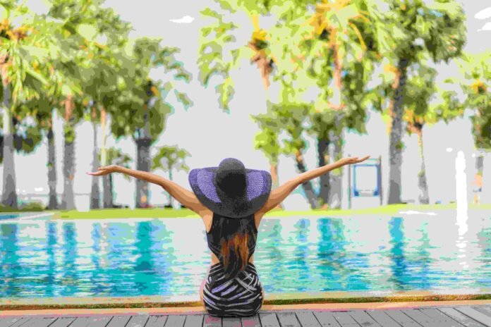 Hóspede do sexo feminino, de costas e braços abertos, está sentada em frente a uma piscina cercada por palmeiras, simbolizando a entrega sensorial e a conexão emocional que definem o turismo sinestésico.