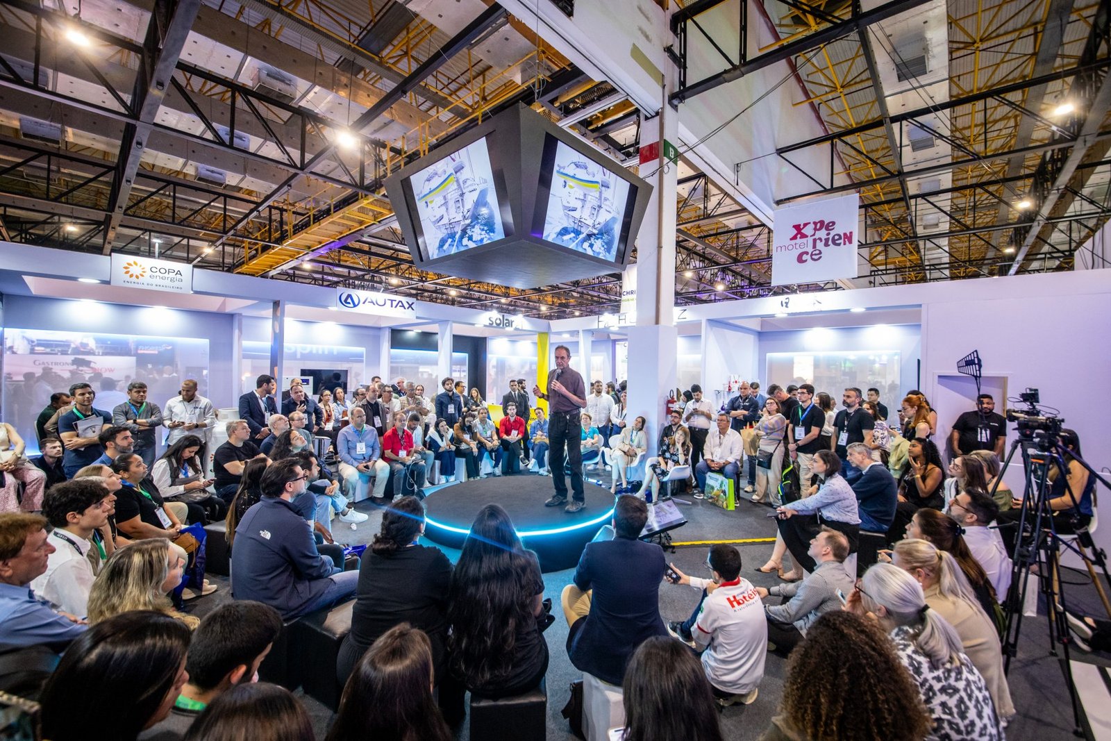 Palestra no espaço Motel Xperience, um dos destaques da programação da Equipotel. Um homem fala de pé em um palco circular com iluminação em neon azul, cercado por uma plateia atenta de profissionais sentados e em pé. Ao fundo, banners do evento e telões suspensos completam o ambiente de inovação e conteúdo do pavilhão de exposições.