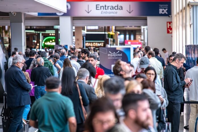Fotografia em plano médio e horizontal de um grande grupo de pessoas reunidas no saguão de entrada da feira Equipotel. A multidão, vista de costas e de perfil, movimenta-se em direção ao acesso do evento, sob uma placa azul com setas e o texto 